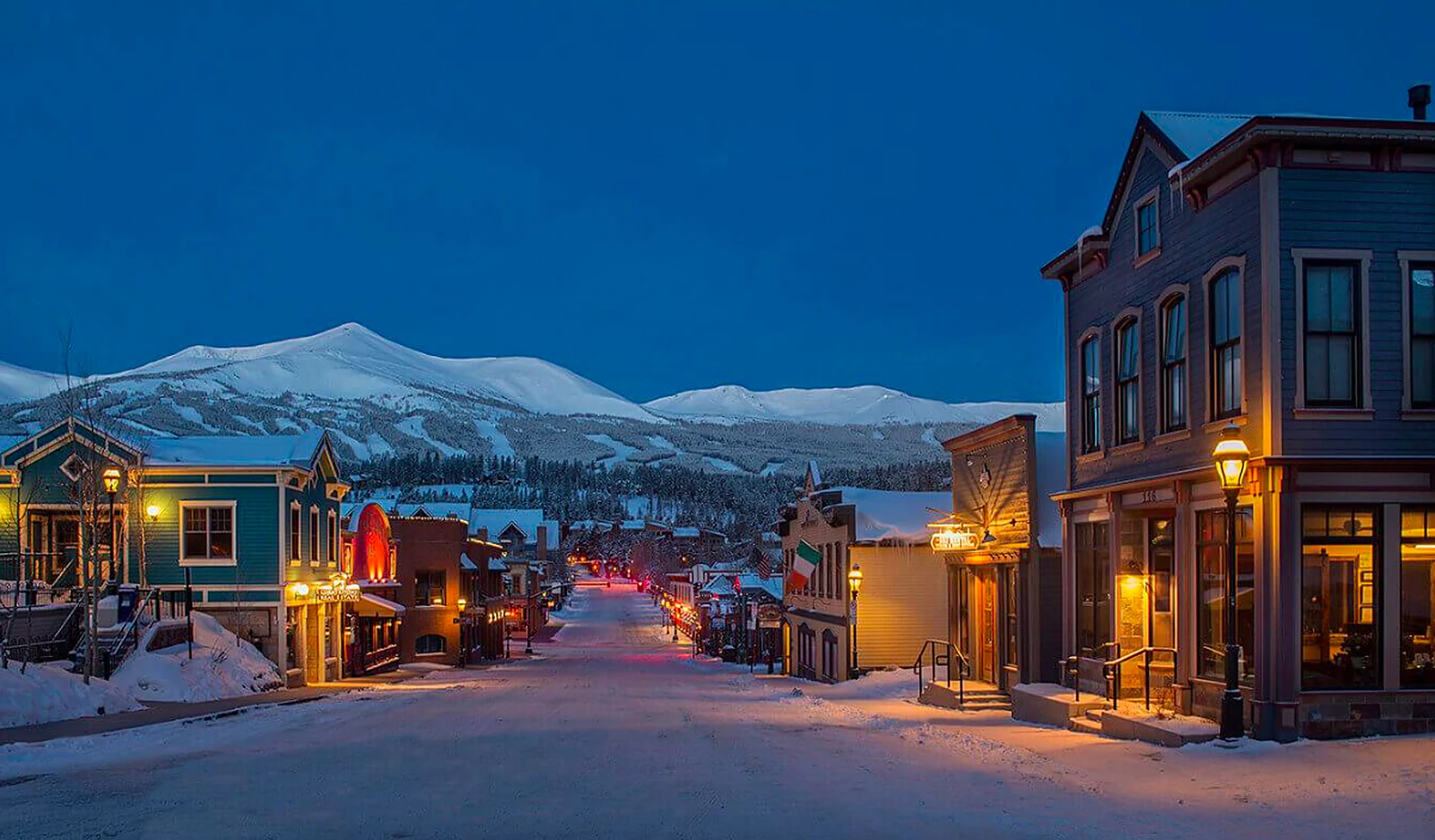 Historic Main Street in Breckenridge, Colorado, glowing at winter dusk with snow-covered peaks rising behind the Victorian storefronts